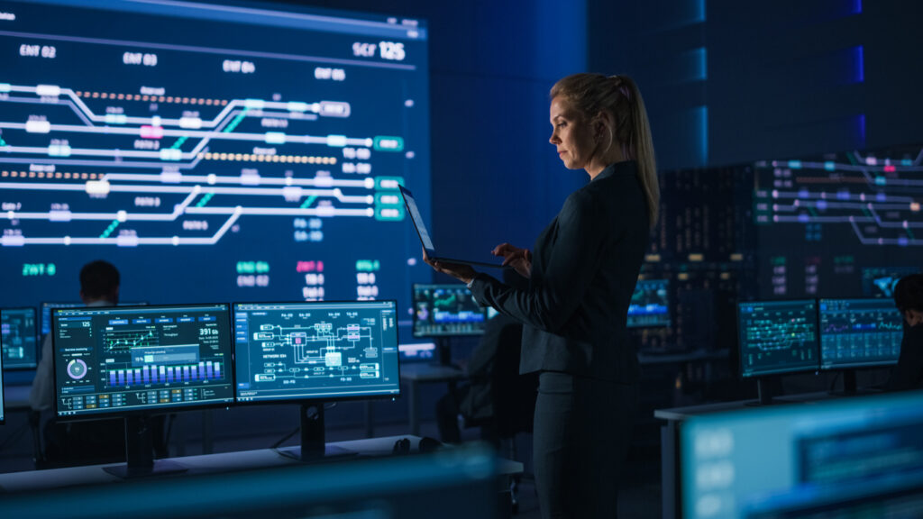 Beautiful Confident Female Project Manager works on a Laptop Standing in Telecommunications System Control Room. In Background Screen Showing Infographics of Infrastructure, Charts, System Analysis